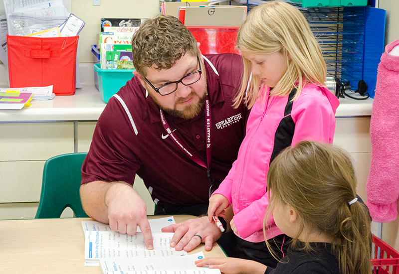 A male teacher helps two female elementary students in a classroom.