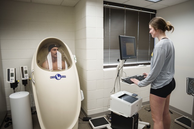 A BOD POD body composition measurement device in an Exercise Science lab. A student sits inside the egg-shaped white pod chamber with a clear window, while a female technician in a gray shirt operates the connected computer system nearby.