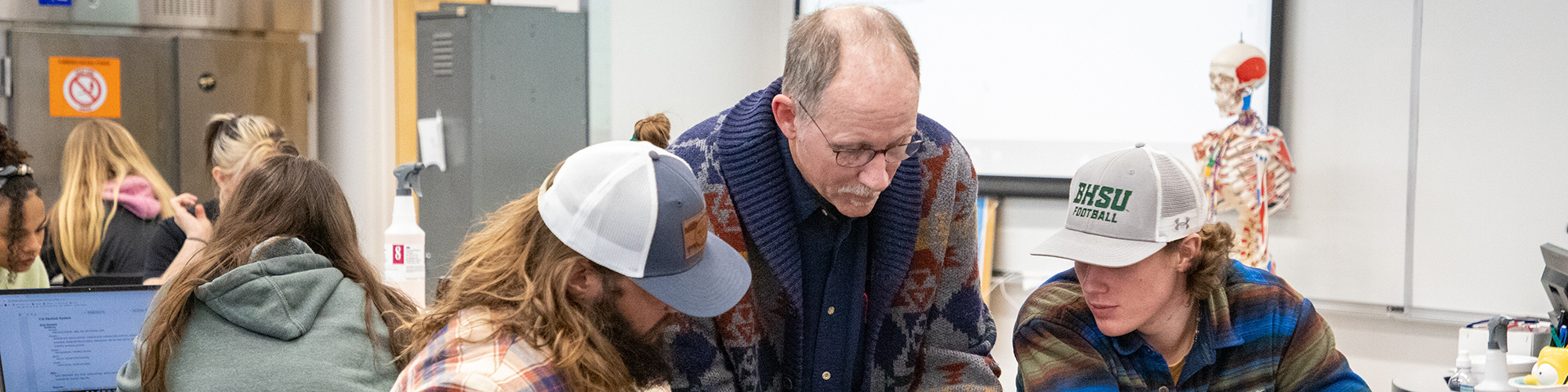 Biology Professor helping two students with hats on in a biology lab and a skeleton model in the background