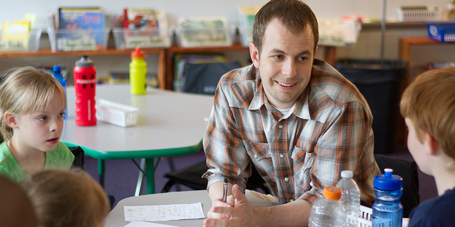 A teacher sits with elementary school students in a classroom.