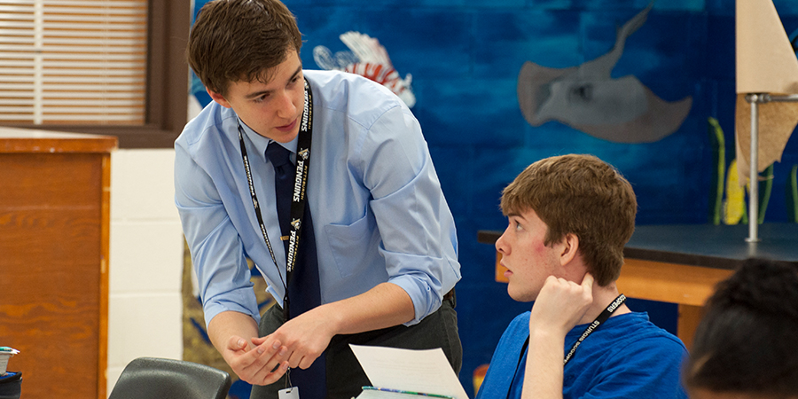 Two men having a conversation. The man on the left is standing and the man on the right is sitting.