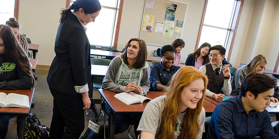 Nine students sit in a classroom while a professor talks to one more student.