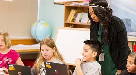 A women watches four elementary students use laptop.