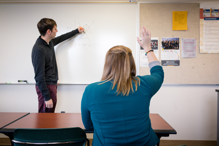 A woman raises her hand as a man writes on a whiteboard in the background.