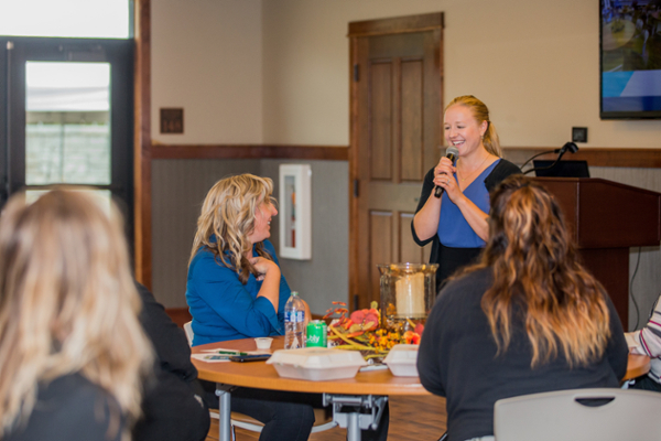A woman stands talking into a microphone in front of a group of sitting women.