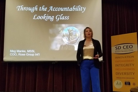 A woman stands on stage with a PowerPoint behind her on a slide that reads, "Through the Accountability Looking Glass."