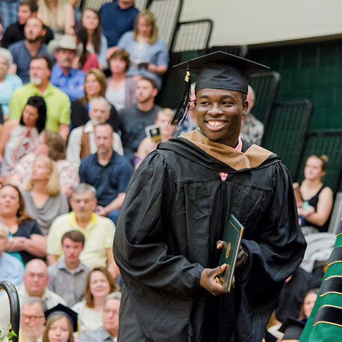 Picture of BHSU graduate accepting his diploma; large audience in the background sitting in the bleachers of the Young Center Gym