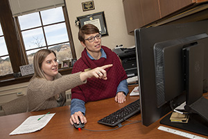 Two women sitting at a desk, one woman pointing at computer other woman looking at computer