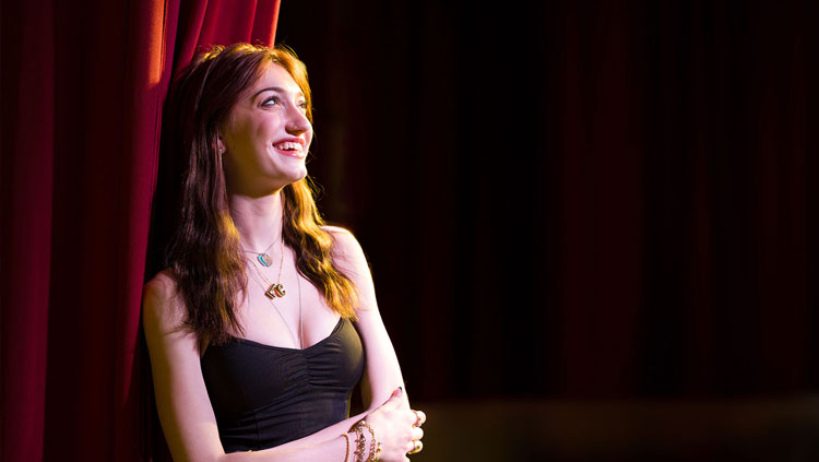 BHSU student Logan Culhane leans against a red curtain on the stage in the Woodburn Auditorium. She is wearing a black dress and smiling.