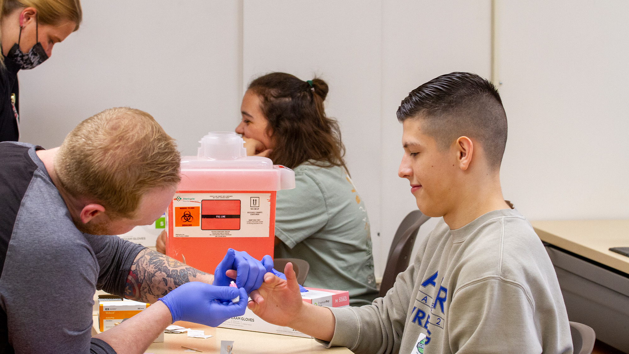 A scrubs camp camper has his finger pricked by a health care worker while learning proper blood drawing procedures and safety in health care