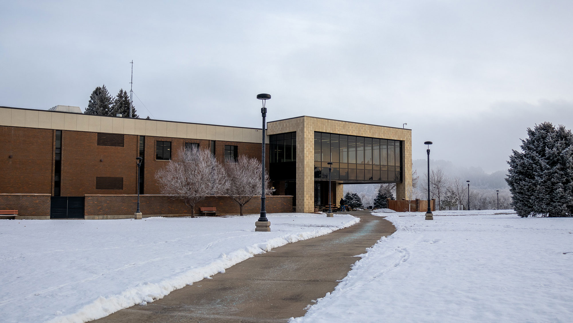 Winter scene at Black Hills State University with snow-covered ground and a curved pathway leading to the E.Y. Berry Library with large glass windows and brick walls.