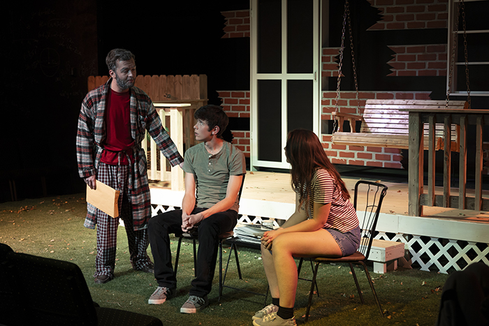 One man is standing and looking at a guy and a girl sitting in two chairs and facing him.