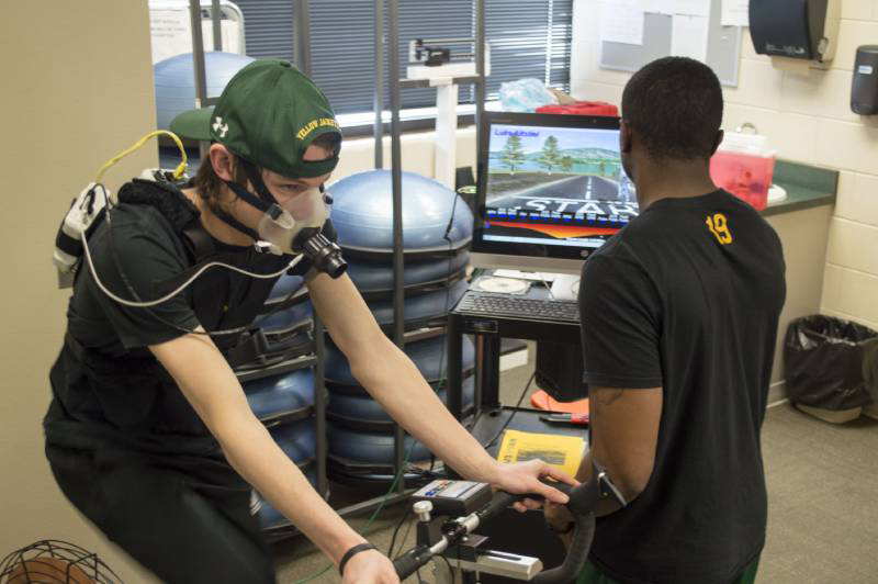 Student on the left rides an exercise bike while the student on the right monitors a screen.