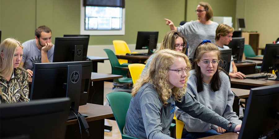 Six students sit at computers while a professor stands in the back of the room.