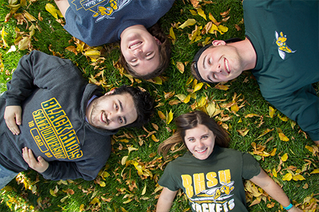 Four students lay in a circle with their heads together pose for an overhead photograph.