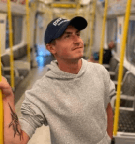 A young man with short brown hair and a backwards baseball cap stands on a train. He has a tattoo on his forearm and is looking up and to the side, away from the camera.