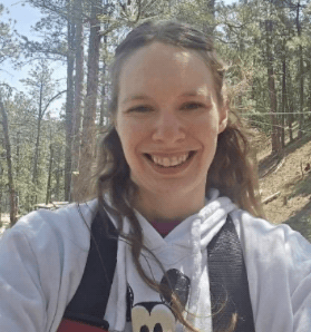 A woman with long brown hair stands in a forest of tall pine trees, smiling directly at the camera. She's wearing a white hoodie with a black Mickey Mouse outline on the front.