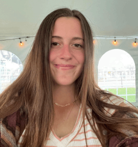 A young woman with long brown hair smiles at the camera, with a white tent structure in the background. She is wearing a brown and white plaid flannel over a light-colored shirt and a gold chain necklace.
