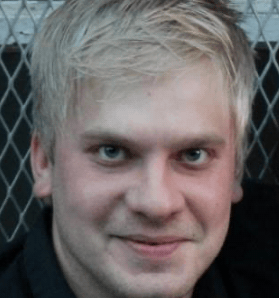 A man with short blond hair looks straight at the camera with a neutral expression. He is wearing a dark, collared shirt