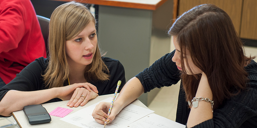 Woman on the left converses with a woman who is writing in a notebook.