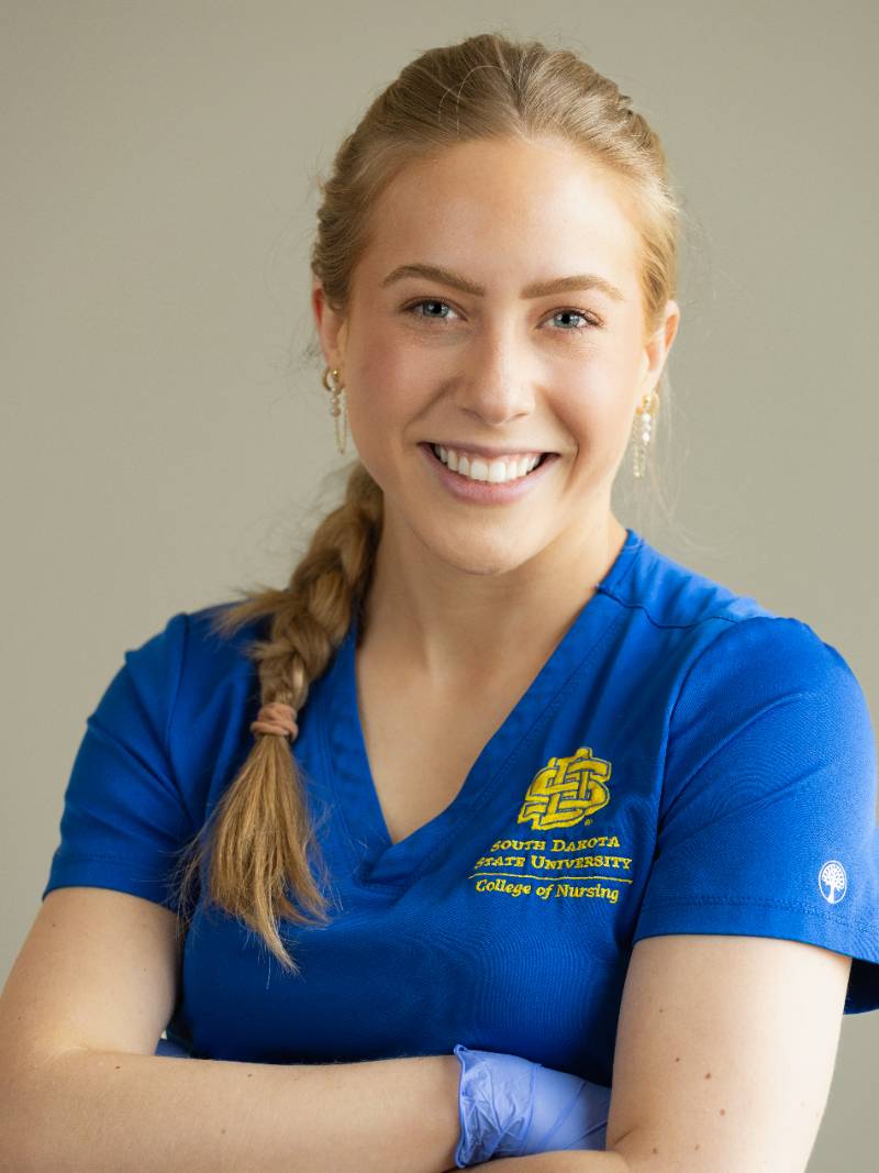 Person in blue SDSU nursing scrubs with braided hair and earrings, smiling at camera with arms crossed against a neutral background.