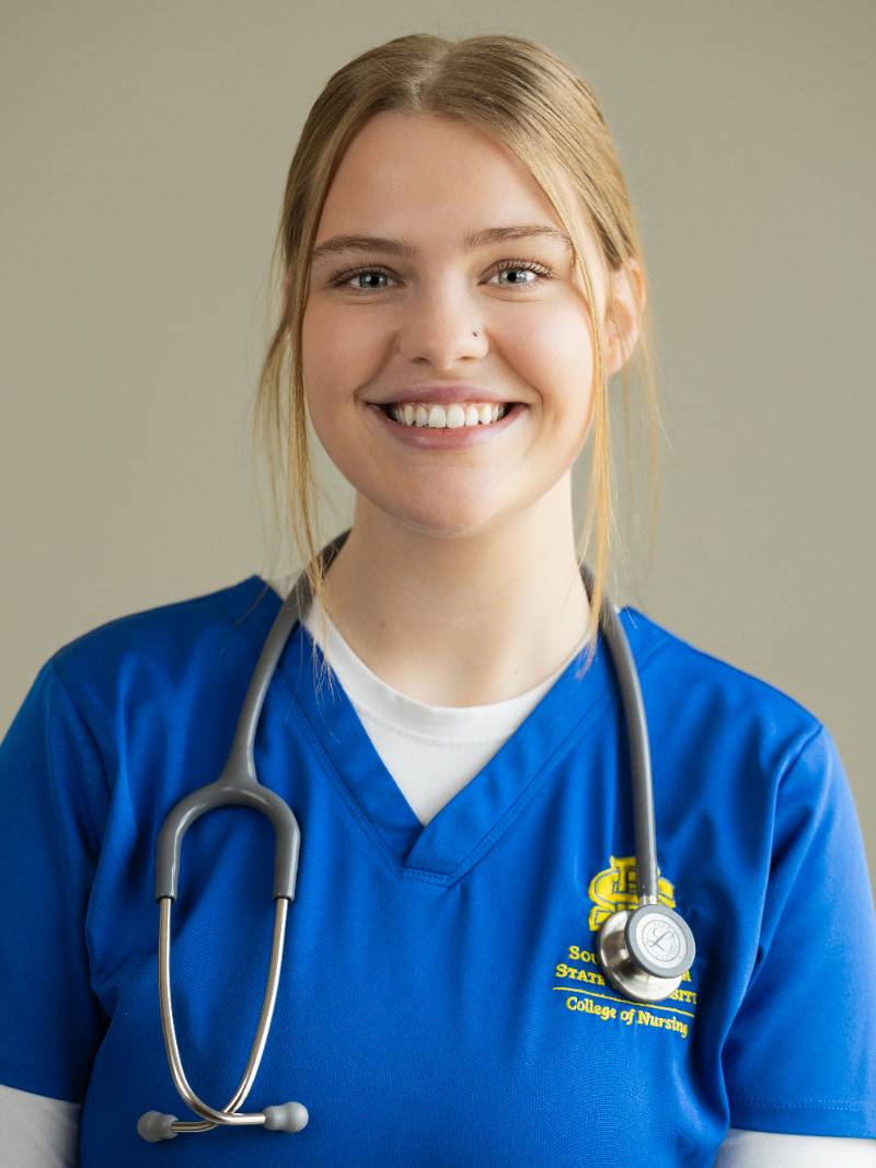 Person in blue SDSU nursing scrubs with a stethoscope, smiling at camera against a neutral background, hair pulled back.