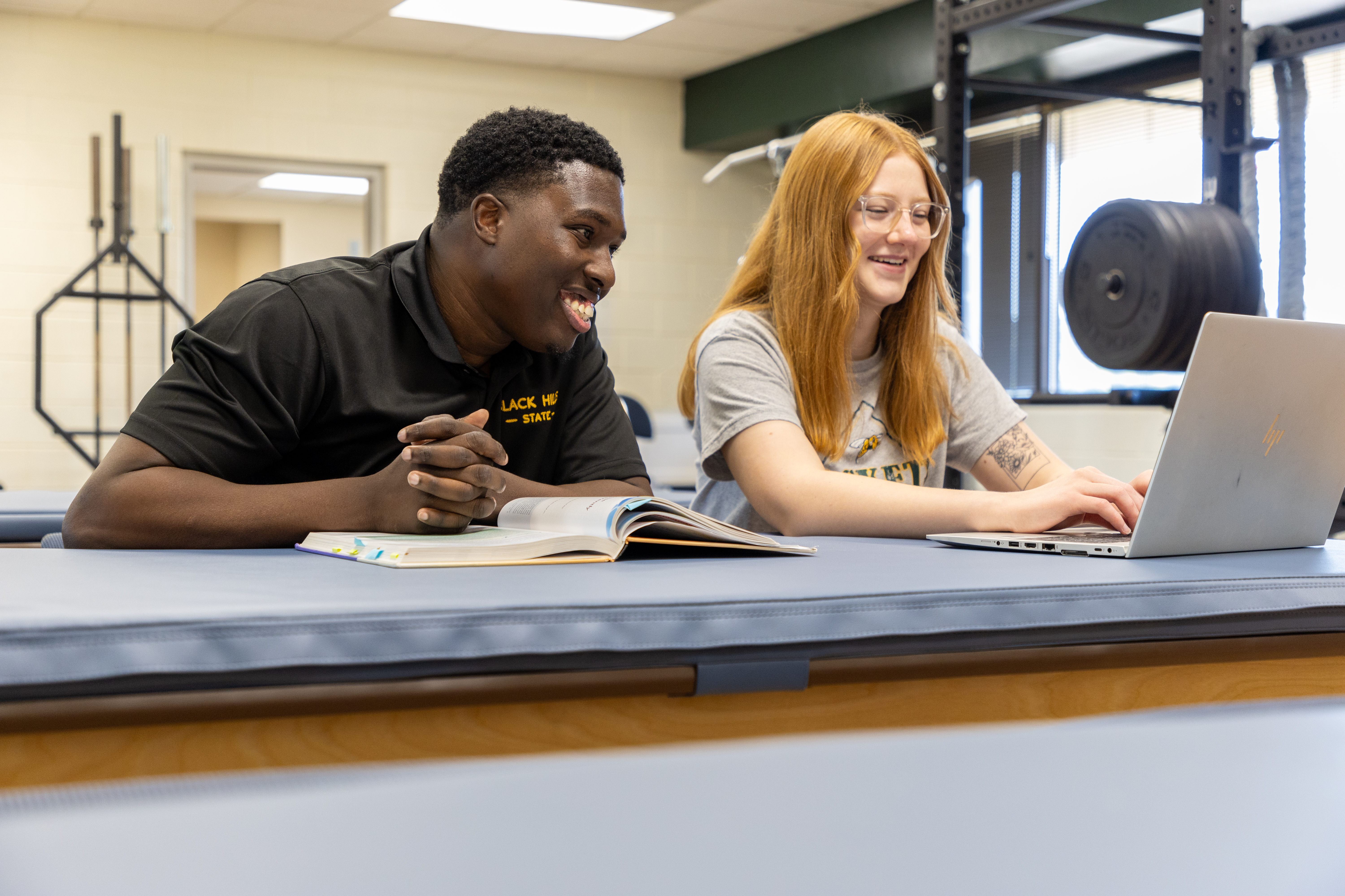 A BHSU professor and student sit together in a gym. The professor smiles while watching the student type on a laptop, with an open textbook in front of them.