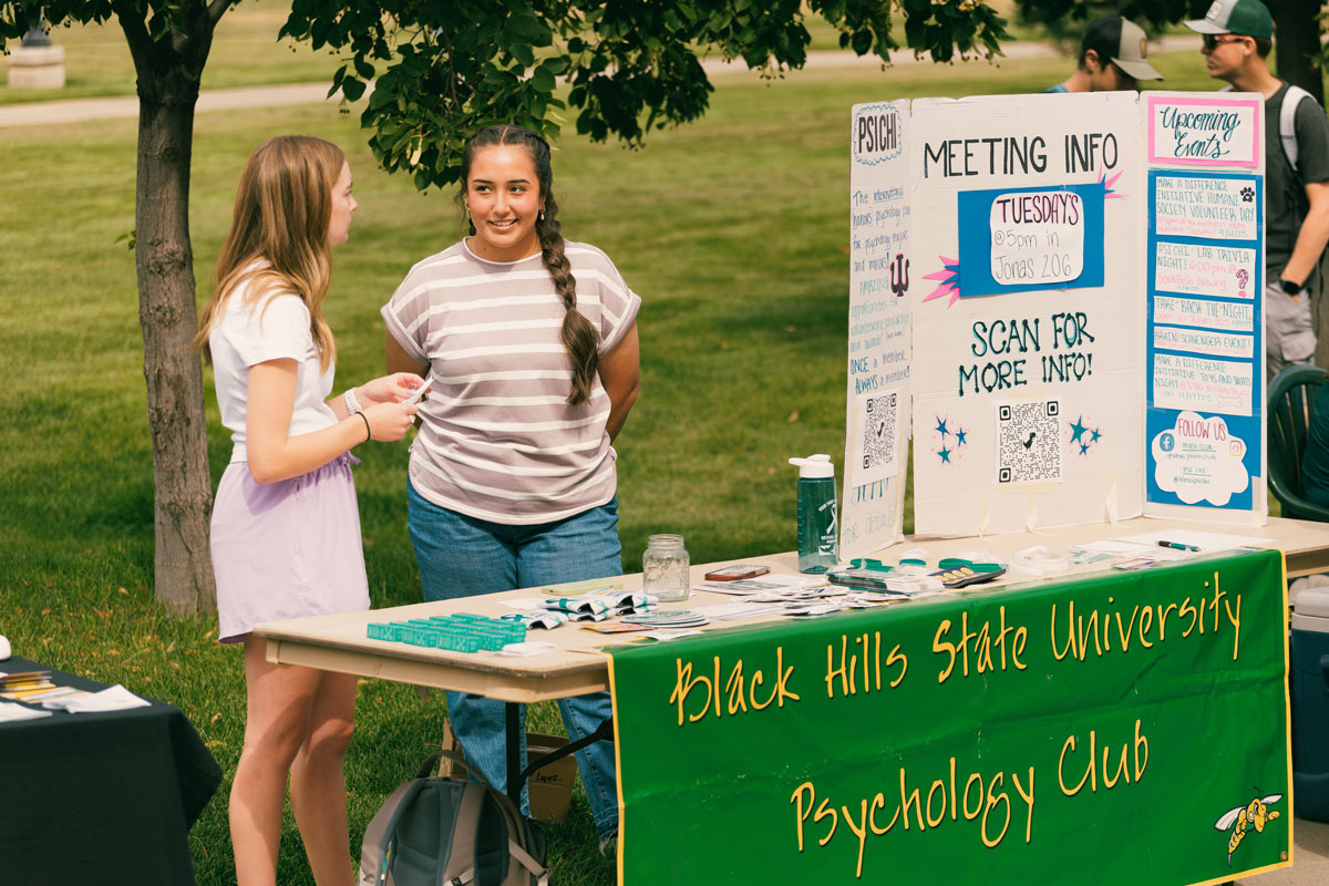 two females studnets stand behind a table on a sunny day. The banner on the table says "Psychology Club."