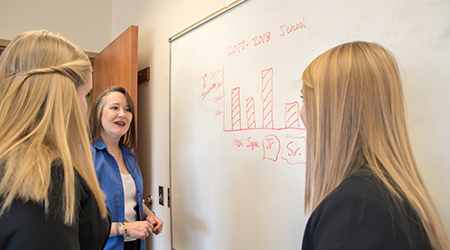 Three women speak to each other in front of a whiteboard.