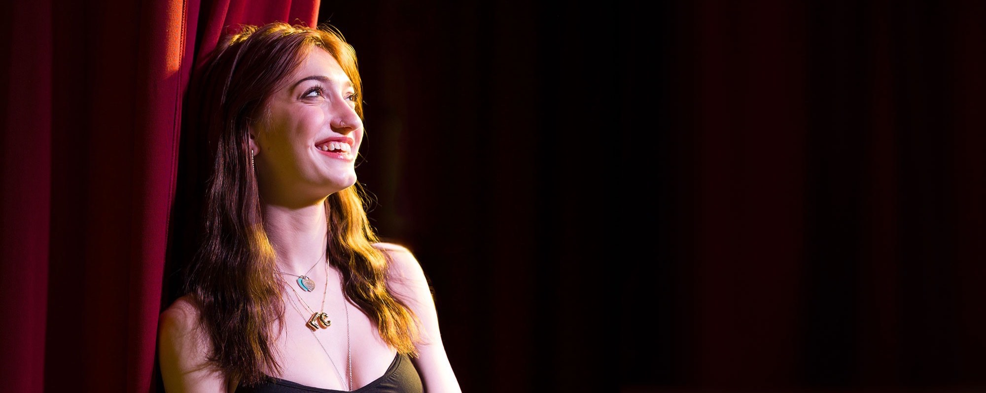 BHSU student Logan Culhane leans against a red curtain on the stage in the Woodburn Auditorium. She is wearing a black dress and smiling.