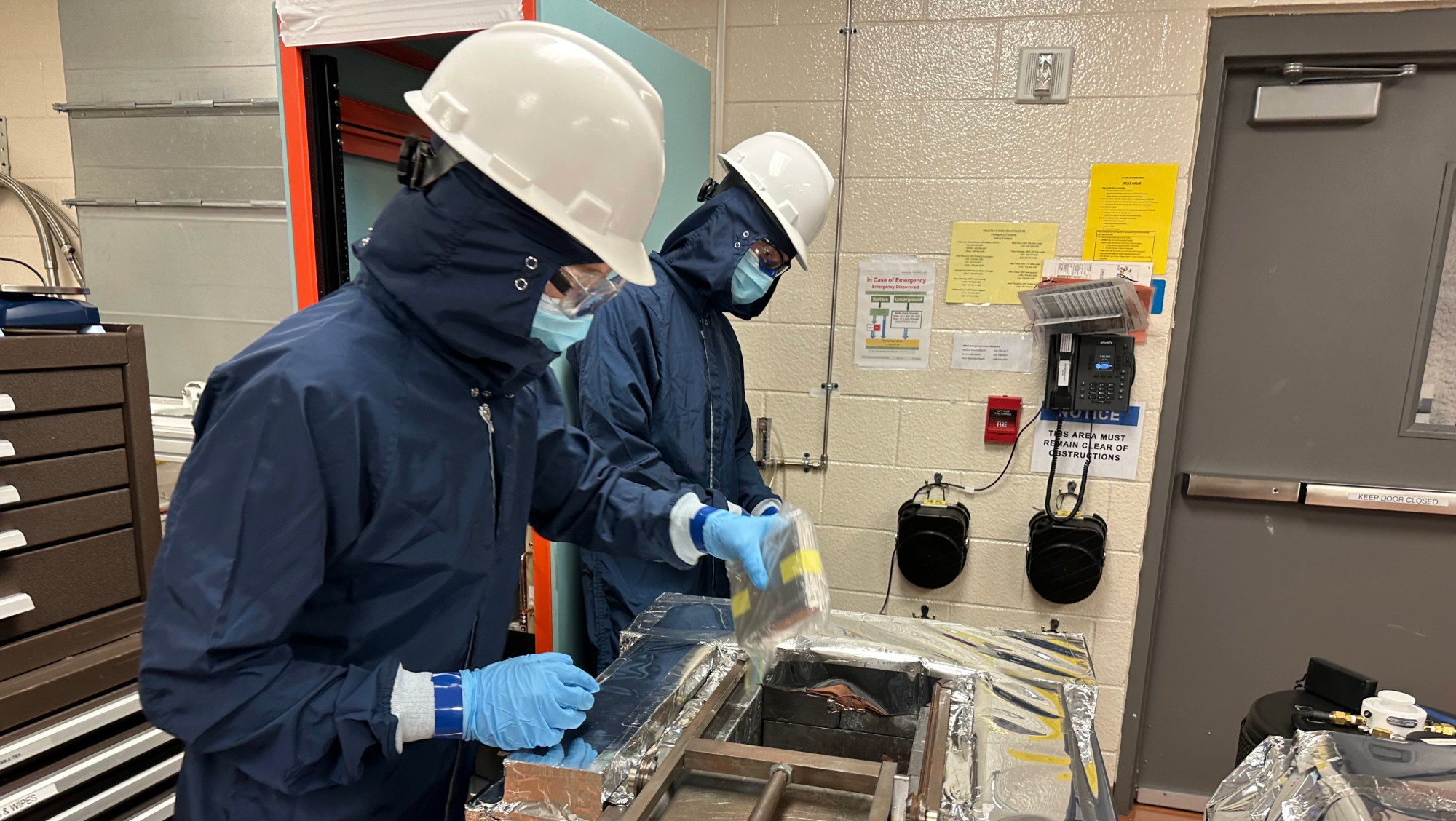Two researchers in protective gear work with equipment in a lab, one holding a sealed bag over a foil-covered machine.