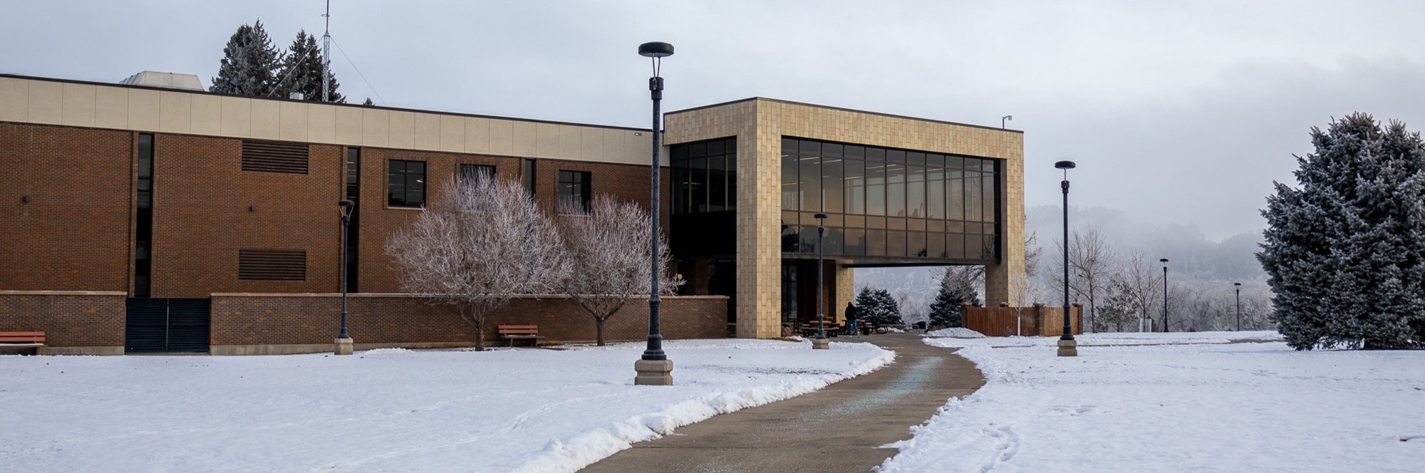 Winter scene at Black Hills State University with snow-covered ground and a curved pathway leading to the E.Y. Berry Library with large glass windows and brick walls.