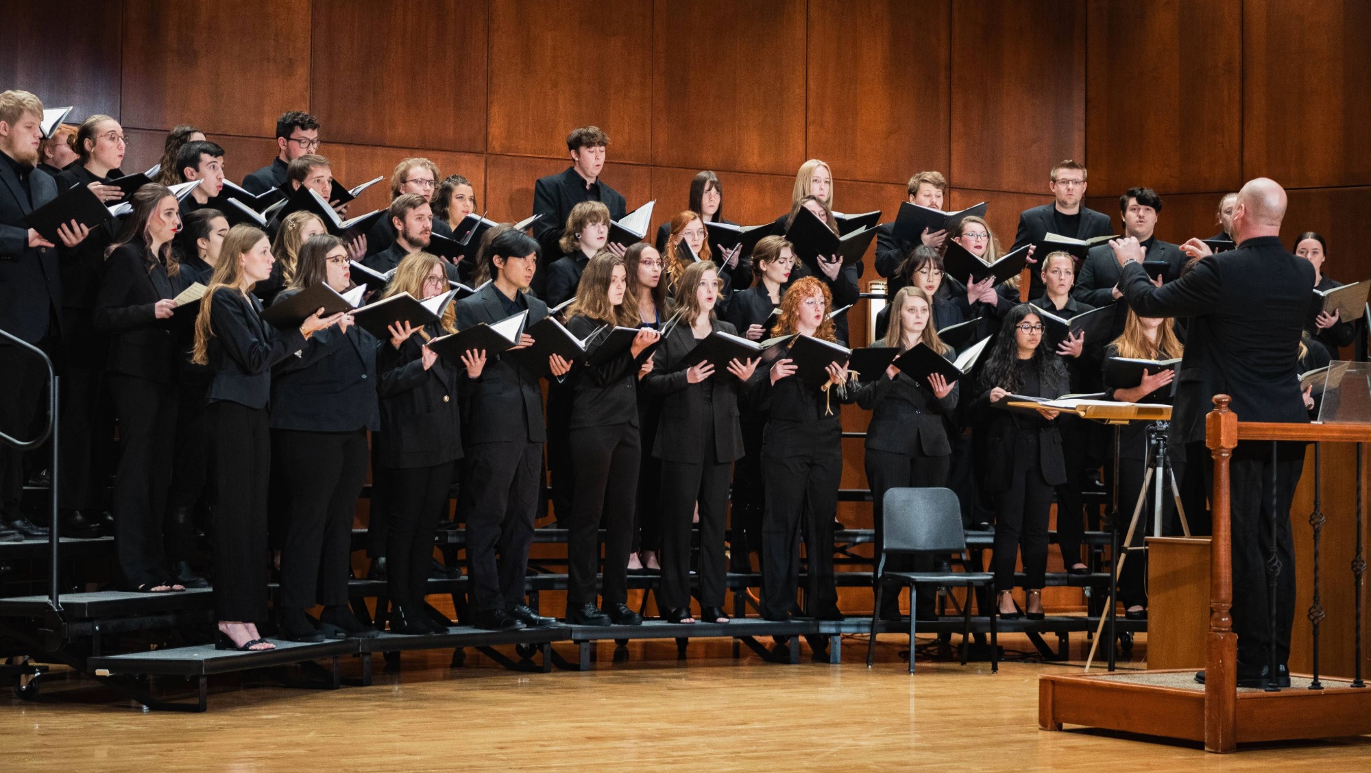 A choir dressed in black sings on stage while a conductor directs them. 