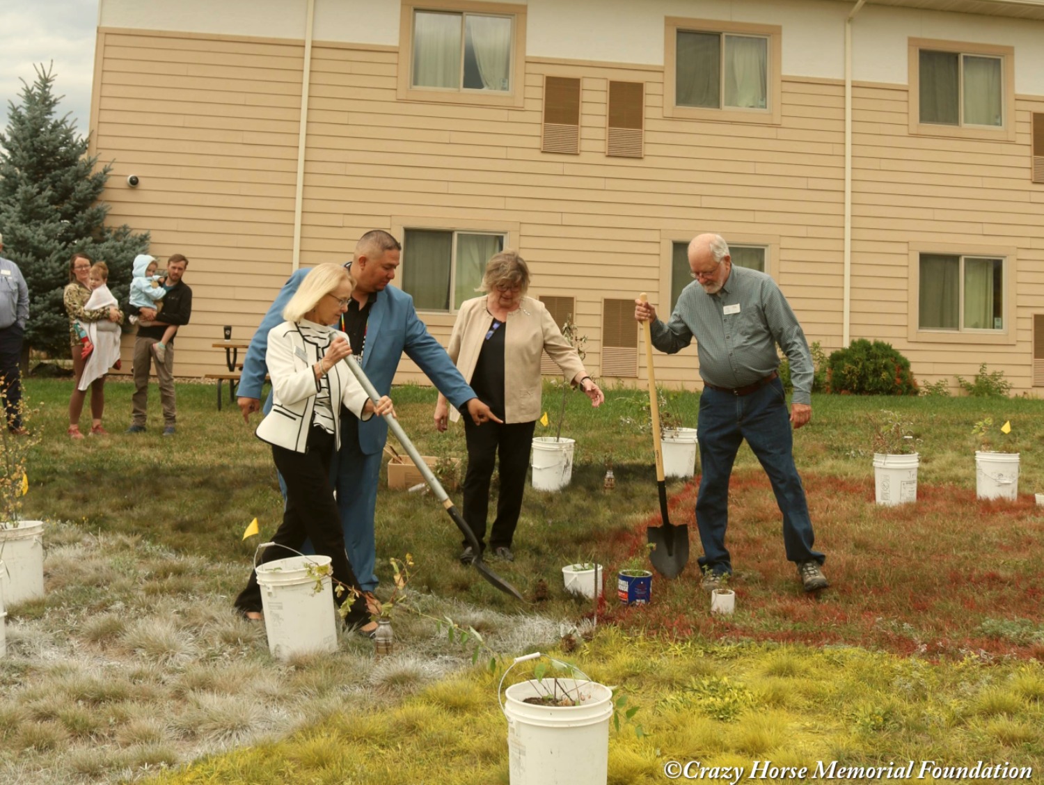 In front of a building, two men and two women hold shovels as they work on the greenhouse project