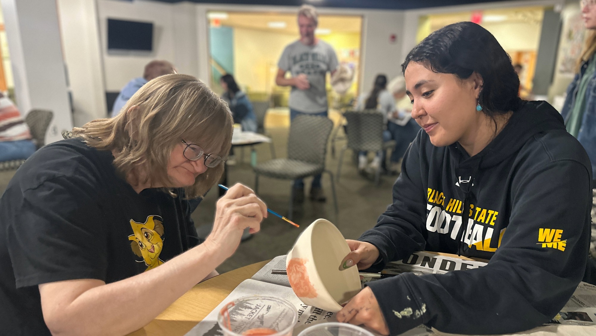 A woman with black hair helps a woman with glasses paint a ceramic bowl