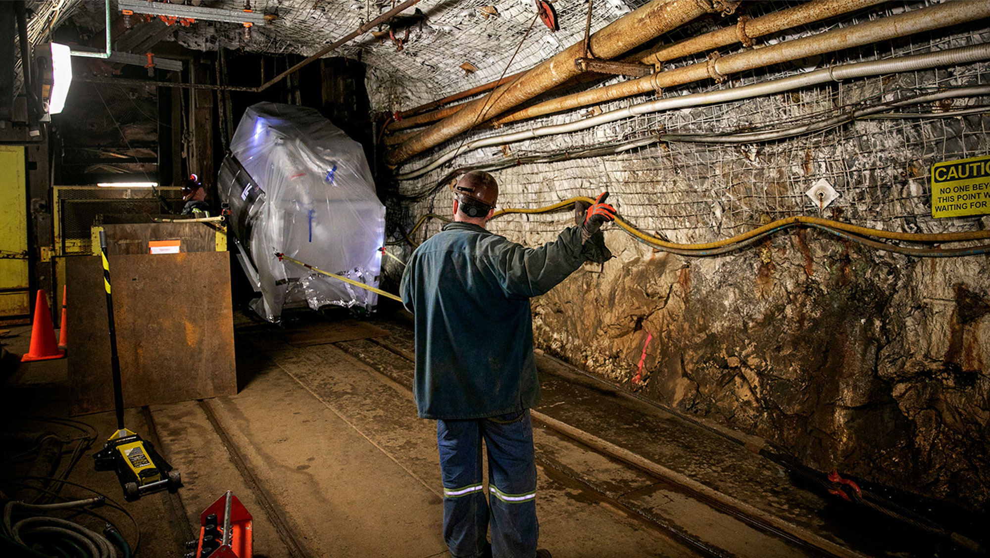 A worker in protective gear maneuvers a large, covered object within a dimly lit underground tunnel. The environment features exposed rocks, pipes, and caution signs, highlighting safety protocols.