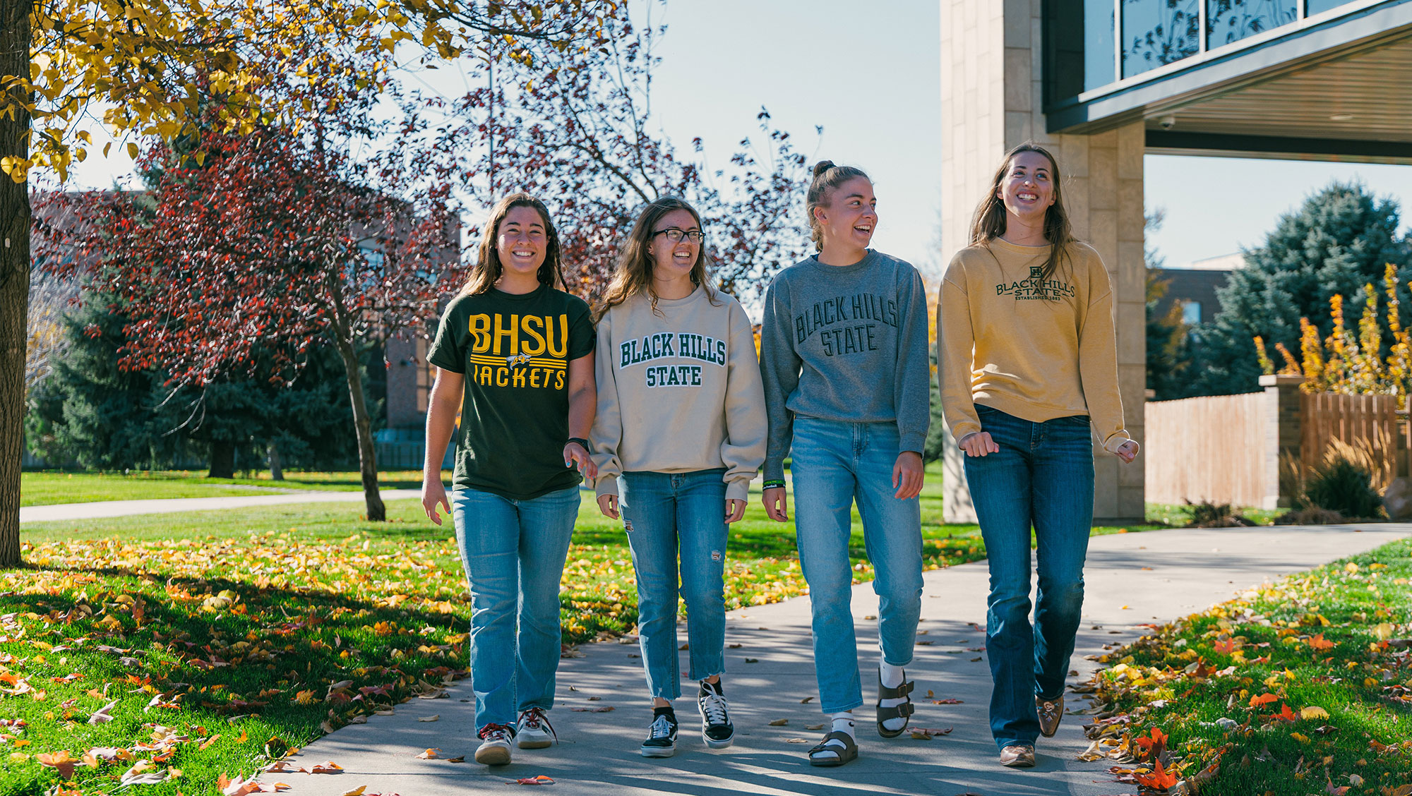 Four BHSU students walk across campus in front of the library in fall. They are smiling and laughing.