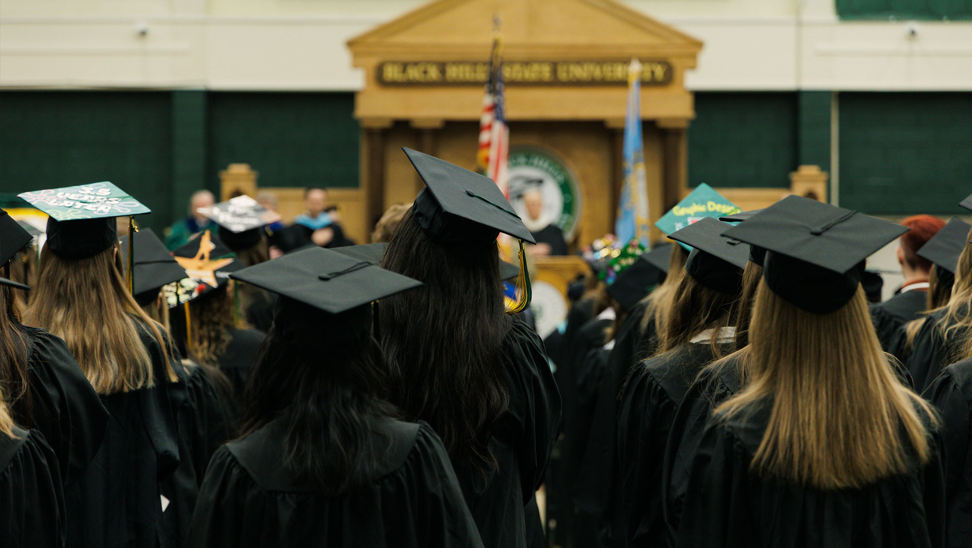 A graduation ceremony at Black Hills State University, showing graduates in black caps and gowns facing a stage with a podium and flags. The background features a wooden structure with the university's name.