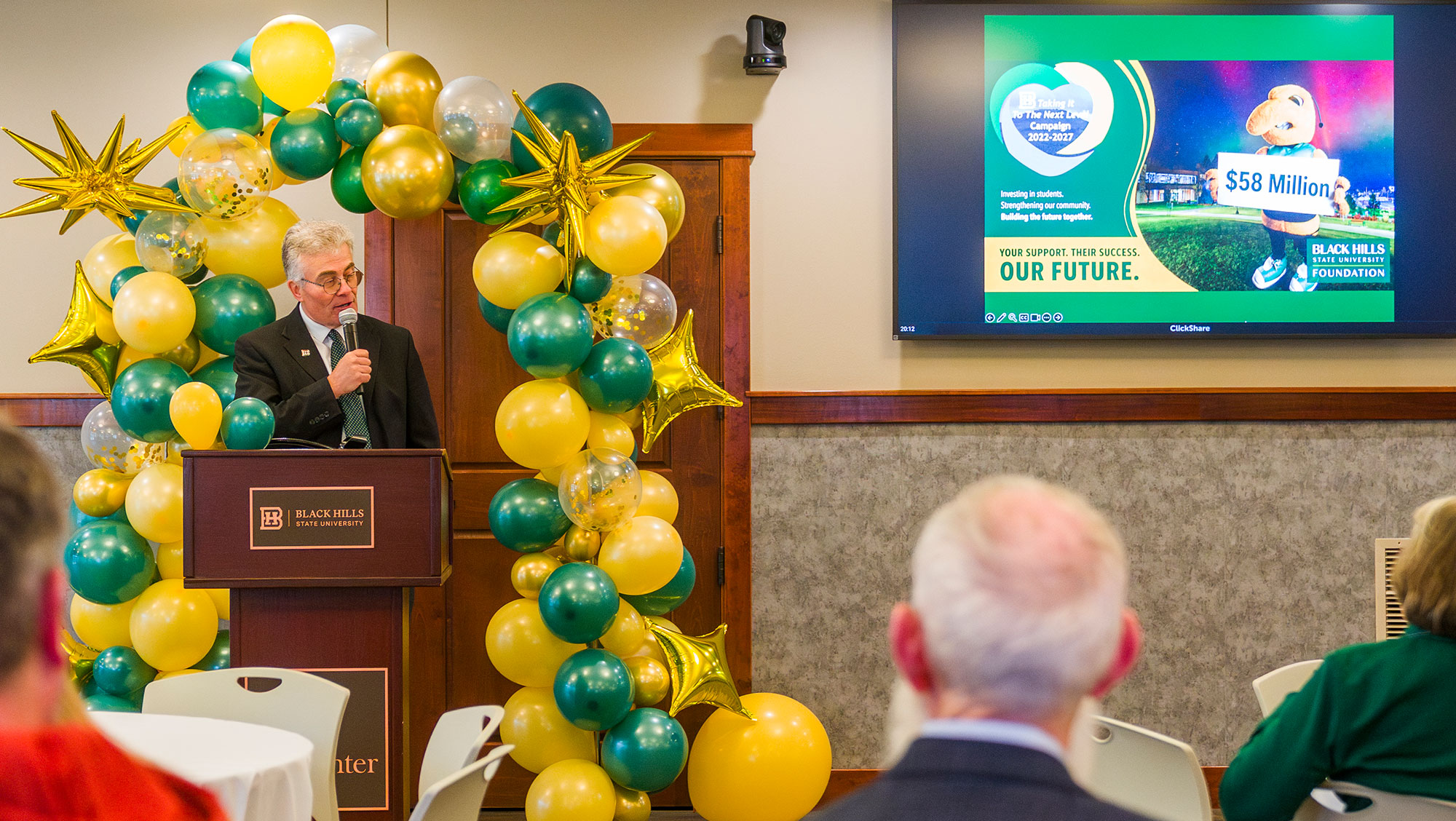 Vice President for Advancement Steve Meeker stands at a podium adorned with a Black Hills State University logo. Beside the podium is a colorful balloon arch with gold, green, and white balloons. A presentation slide on a screen reads 'Helping students, their families, and South Dakota prepare for the future. Your support helps BHSU enhance our future' with a callout of '$58 Million' and logos for Black Hills State University.