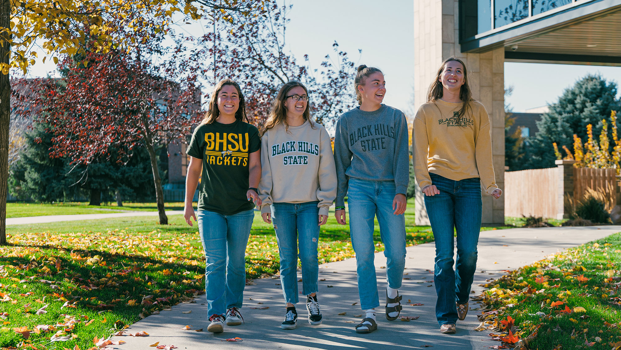 Four BHSU students walk across campus in front of the library in fall. They are smiling and laughing.