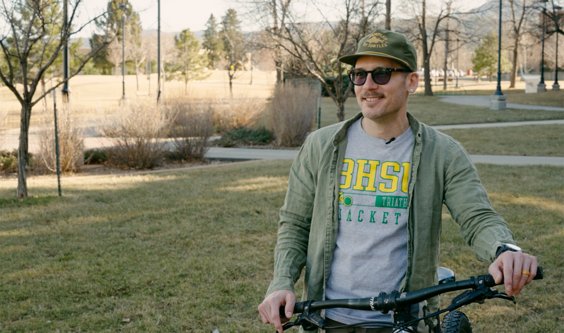 Dr. Trenton Ellis stands with his bike on the BHSU campus. It is a sunny afternoon. He is wearing a hat and sunglasses.