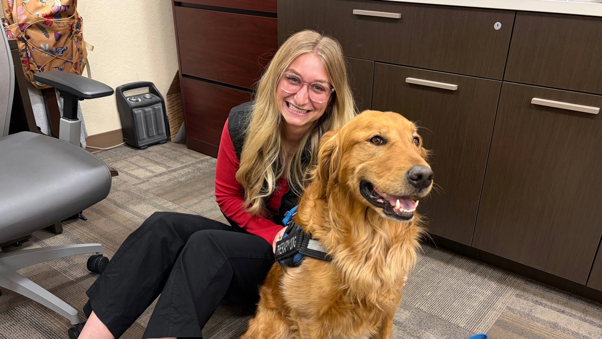 BHSU student Josie Downing sits on the floor with a golden retriever. She has long blonde hair, and glasses. She is smiling. She is wearing a red shirt. The dog has on a working vest.