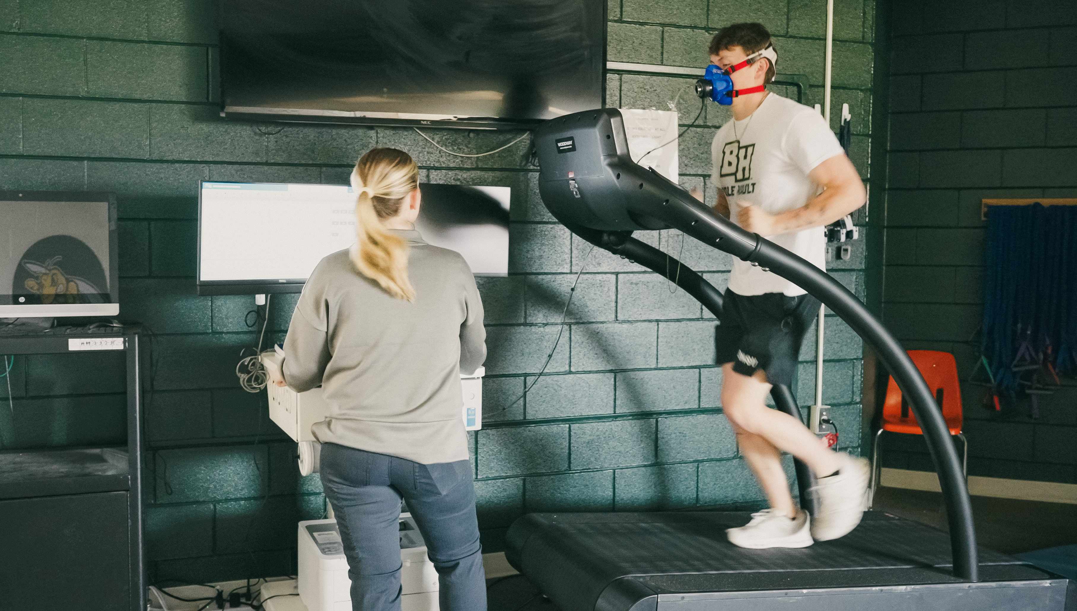 A person runs on a treadmill while wearing a mask for VO₂ max testing as another individual monitors data on a computer in the BHSU exercise science lab.