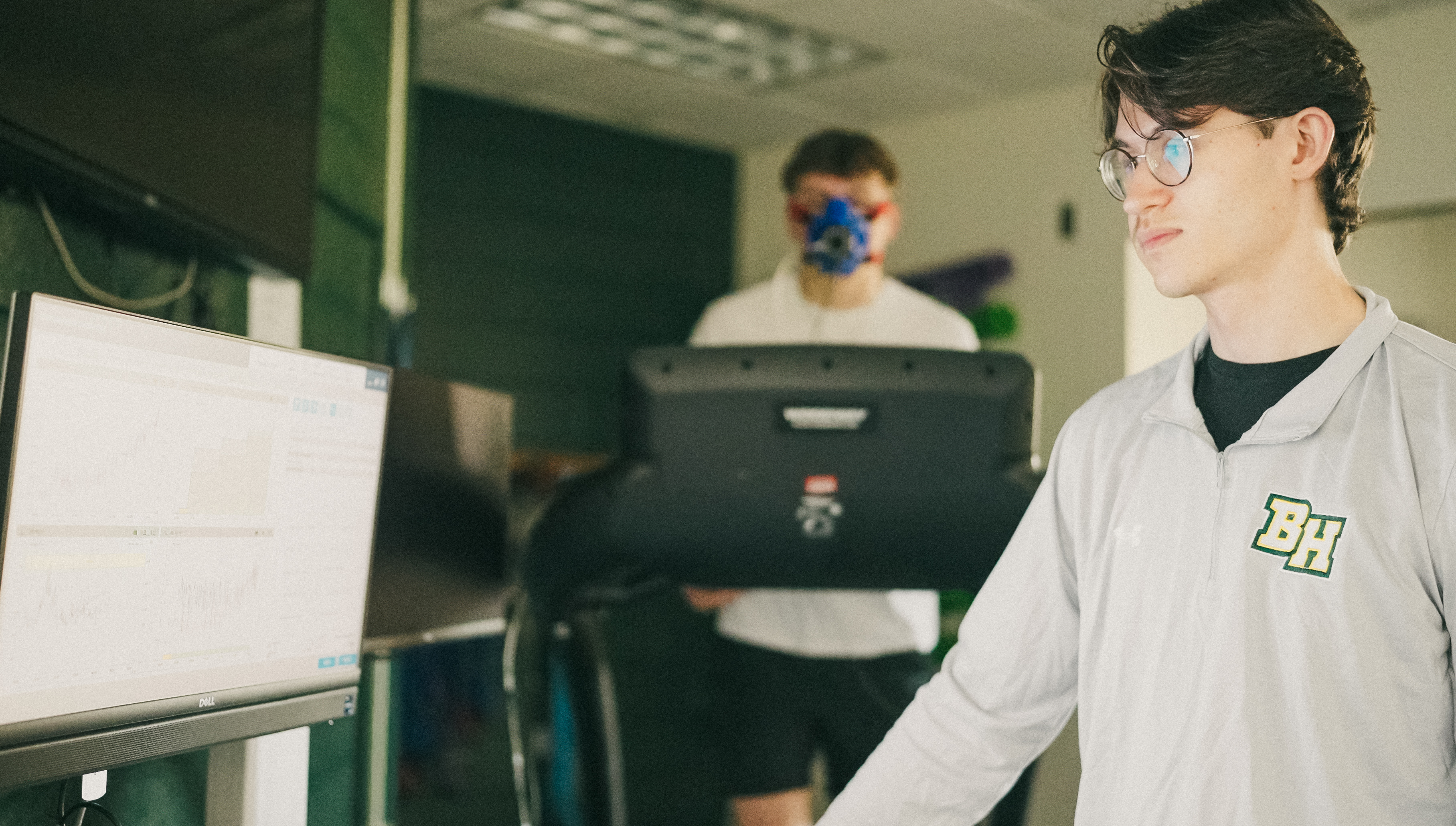 A Black Hills State University Exercise Science student reviews performance testing data in a laboratory while another student participates in physiological assessment equipment in the background.