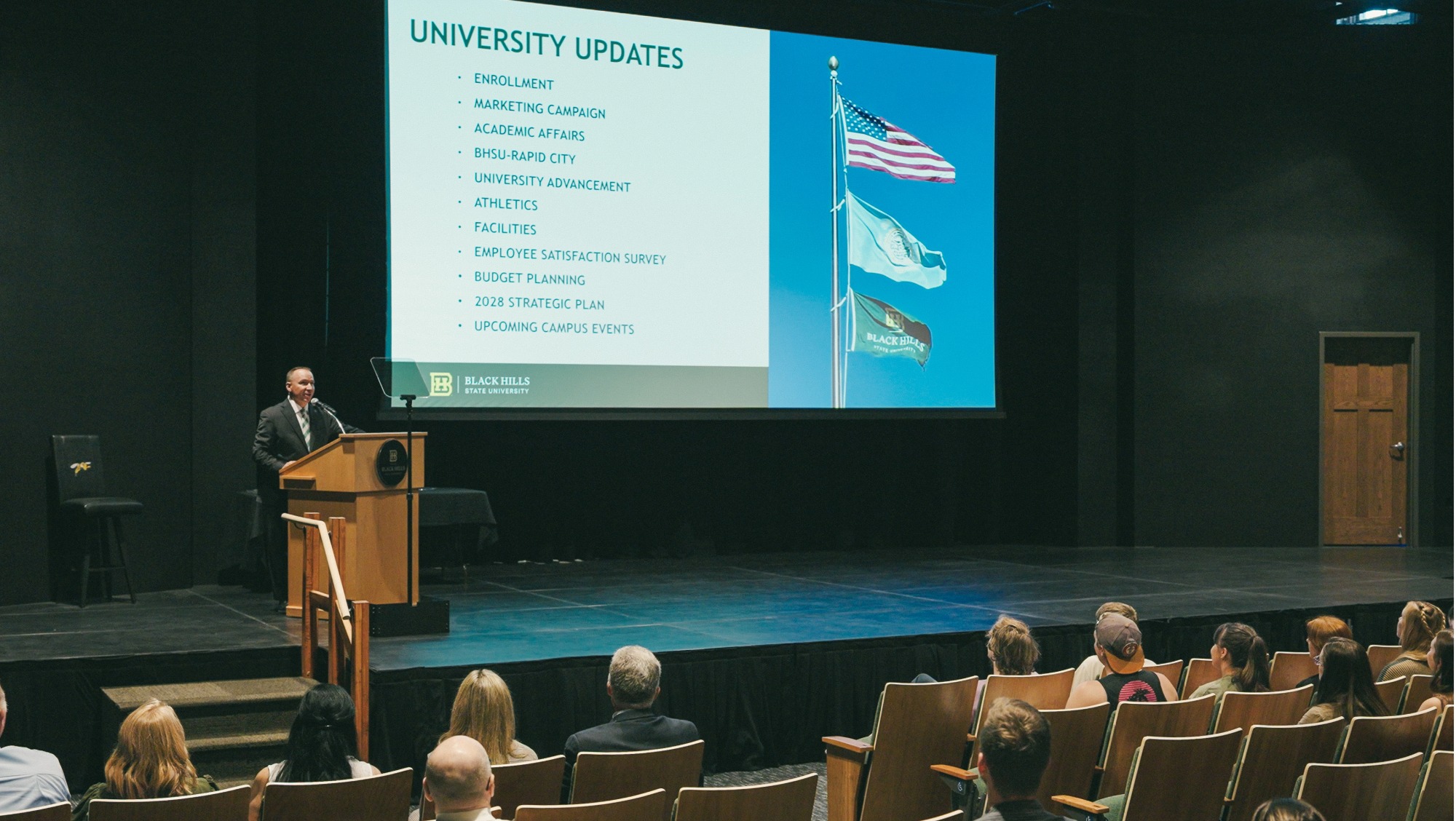 University President Elliot stands at a podium onstage giving a presentation