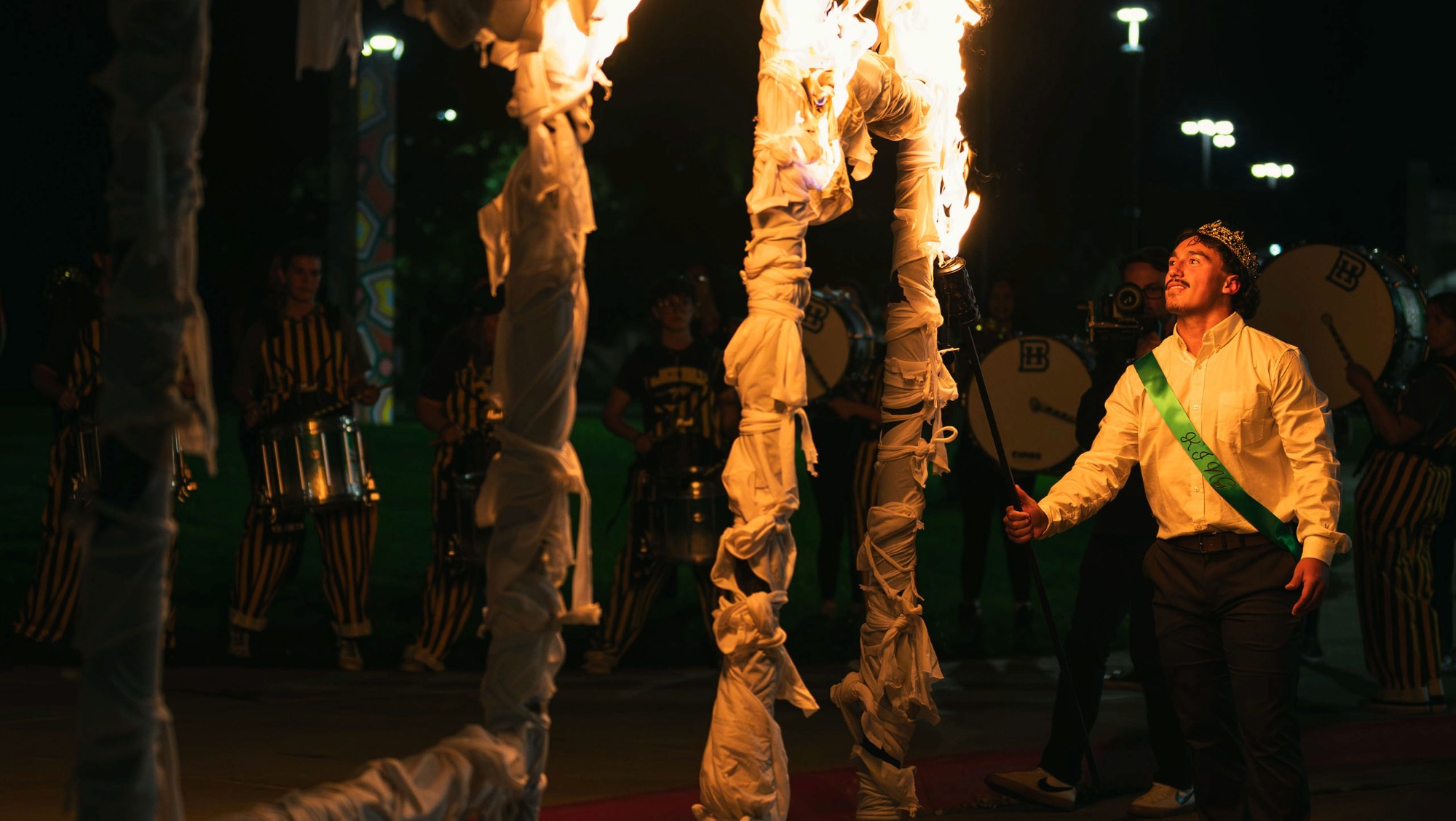 Student wearing a crown and sash lights fire to a large 