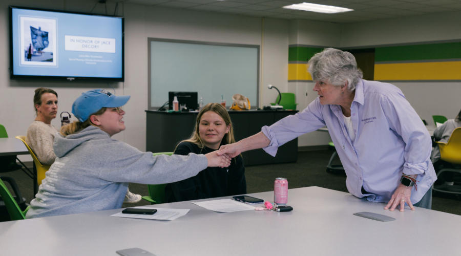 An older woman leans across a table to shake the hand of a younger woman during a lecture taking place in a classroom.