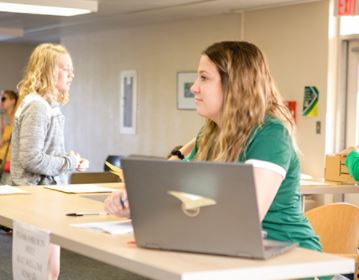 A student sits at a desk with another student walking in the background.