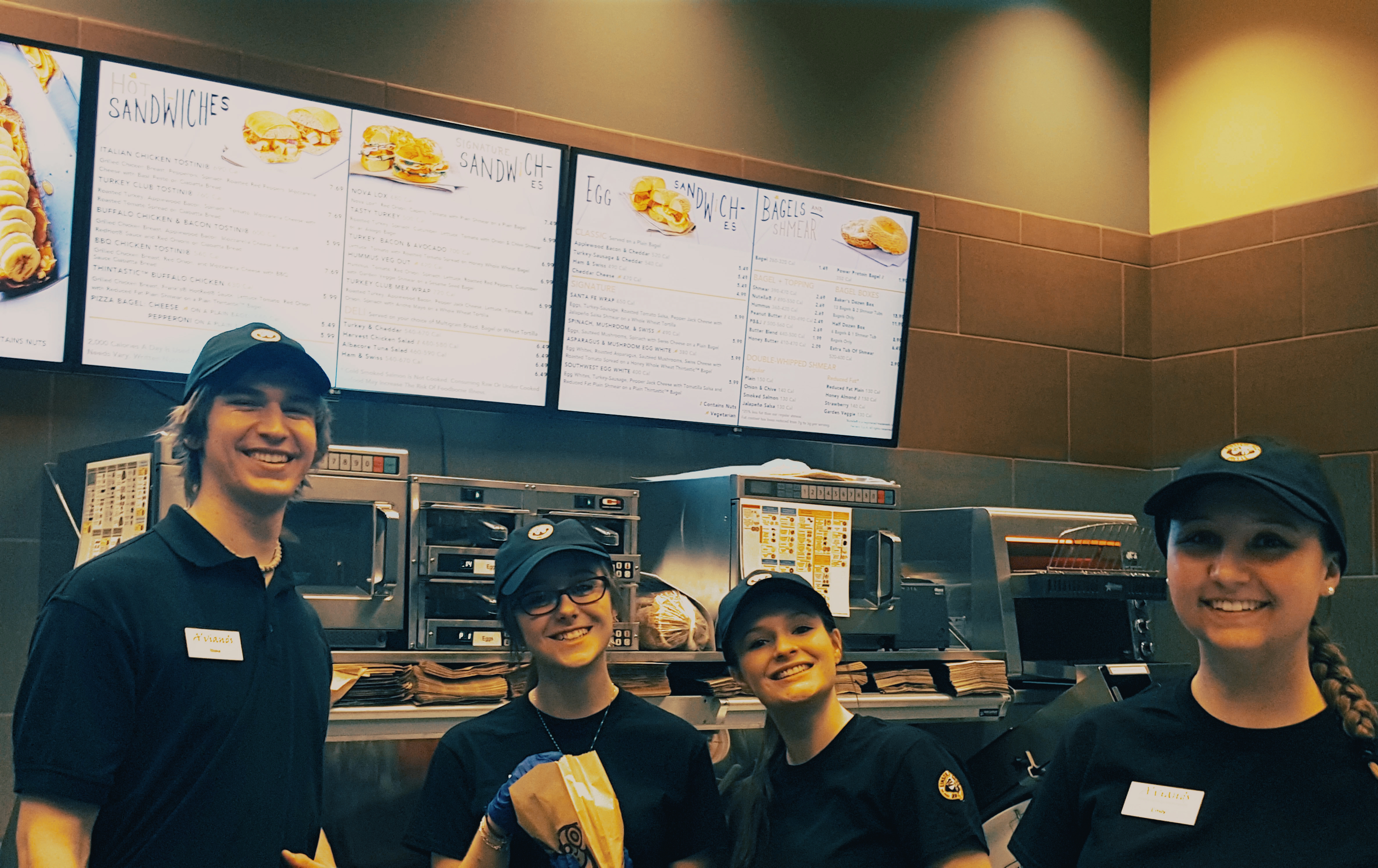 4 Student workers posing in front of the Einstein's menu board. 