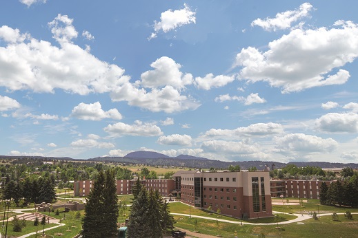 Peaceful college campus featuring modern brick residence halls arranged in a semi-circle, surrounded by lush green lawns and coniferous trees, with rolling hills and mountains in the background under a bright blue sky.